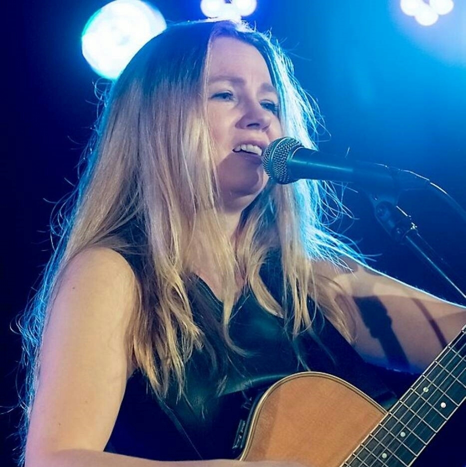 kobieta w długich włosach, z gitarą, śpiewająca do mikrofonu, widoczna od pasa
