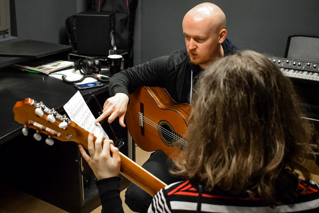 młody mężczyzna siedzący w studio nagraniowym z gitarą, udziela lekcji gry na gitarze młodej osobie