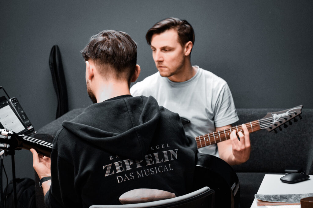 dwóch młodych mężczyzn siedzących w studiu nagraniowym, grających na gitarach, siedzą naprzeciwko siebie, odbywa się lekcja gry na gitarze 