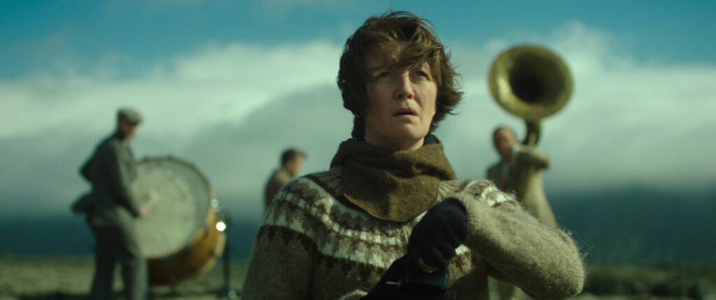 Kadr z filmu "Kobieta idzie na wojnę" przedstawiający kobietę z muzykami i krajobrazem w tle.