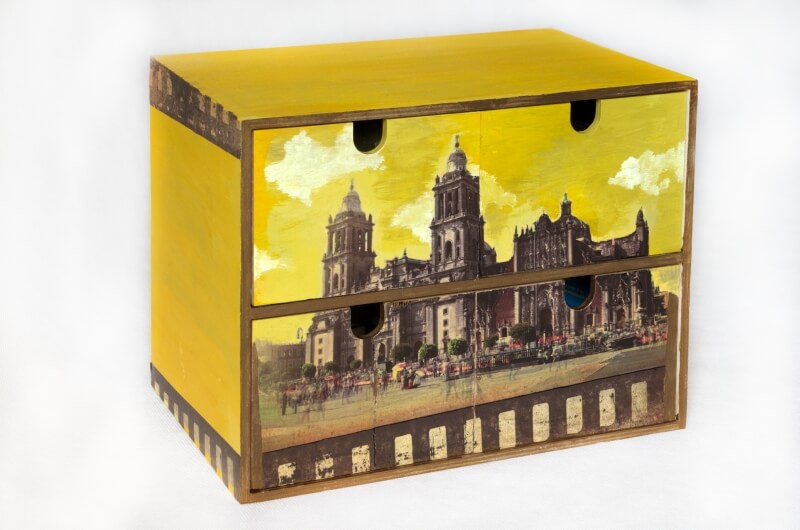 Skrzynka z szufladkami ozdobiona przez Katarzyną Konarską. Na froncie przegród widać zabytkowy budynek oraz niebo w żółtym kolorze.