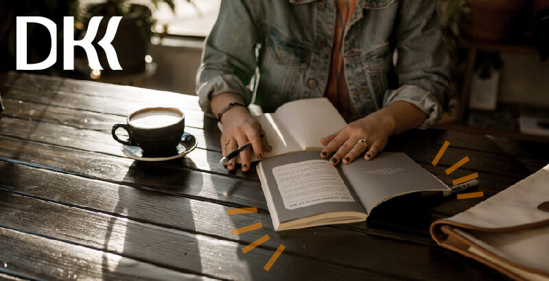 Na zdjęciu widać osobę, która czyta książkę oraz robi notatki z tej lektury w zeszycie. Poza wymienionymi przedmiotami na stola widać napój (prawdopodobnie kawa) oraz teczkę.