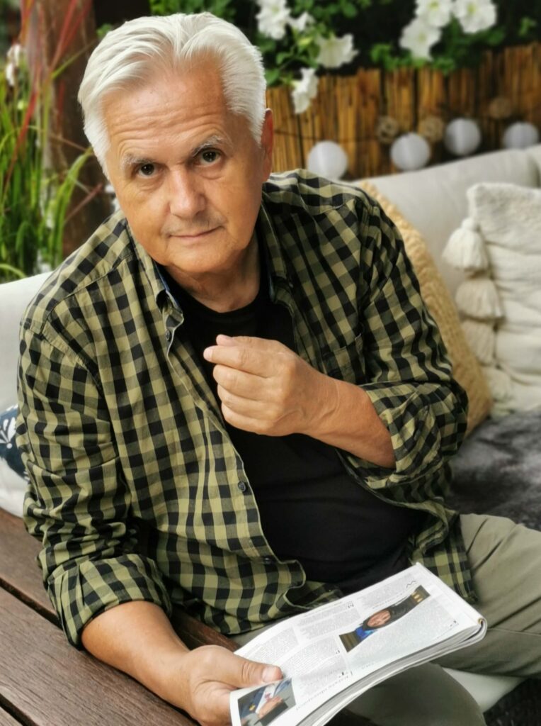 Na zdjęciu widać mężczyznę w wieku około sześćdziesięciu lat, ma siwe włosy, koszulę w kratkę, a w ręce trzyma gazetę.