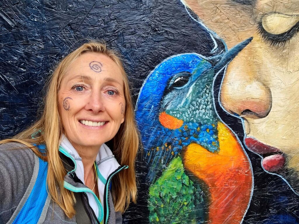 kobieta z tatuażami na twarzy na tle muralu z ptakiem i twarzą
